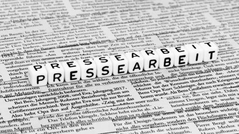 Auf vielen Zeitungsausschnitten mit reinem Text liegen Perlen mit Buchstaben, die das Wort Pressearbeit bilden.