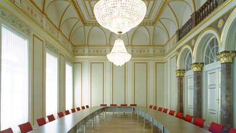 Großer Sitzungssaal Großer Sitzungssaal mit Kronleuchter, Deckengewölbe sowie am Saalrand U-förmig aufgestellte Tische mit Stühlen.