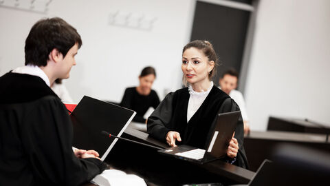 Eine Juristin in Robe zeigt dem Richter an der Richterbank Informationen auf einem Laptop