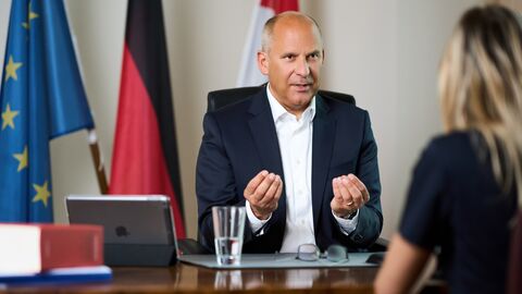 Justizminister Prof. Dr. Roman Poseck während eines Interviews, er sitzt an einem Schreibtisch, ihm gegenüber eine Dame.