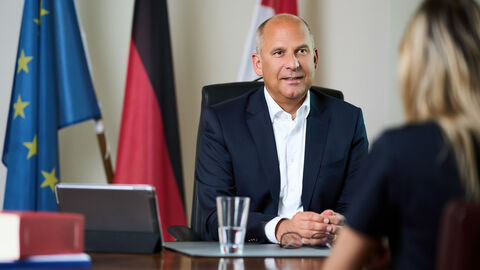 Justizminister Prof. Dr. Roman Poseck sitzt an einem Tisch, im Hintergrund Flagen, im Vordergrund schemenhaft ein Interview-Partner