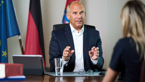 Justizminister Prof. Dr. Roman Poseck sitzt an einem Tisch, im Hintergrund Flagen, im Vordergrund schemenhaft ein Interview-Partner