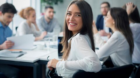 Eine junge Frau in weißer Bluse sitzt auf einem schwarzen Stuhl im Meetingraum und dreht sich lächelnd in die Kamera, im Hintergrund verschwommen weitere Teilnehmer um den Tisch.
