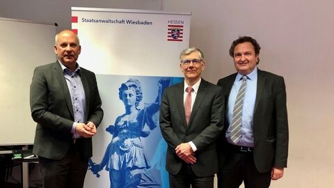 Justizminister Poseck bei seinem Besuch der Staatsanwaltschaft Wiesbaden mit zwei weiteren Personen vor einem Aufsteller mit der Aufschrift "Staatsanwaltschaft Wiesbaden" und einem Bild der Justitia.