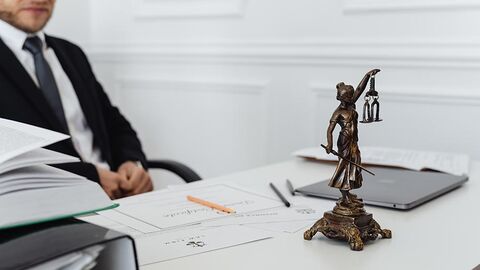 Mann im Anzug sitzt an einem Schreibtisch, auf dem eine Justitia-Figur steht.