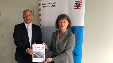 Justizminister Poseck (links) mit Dr. Patricia Wüllner (rechts) vor einem Aufsteller mit der Aufschrift "Amtsanwaltschaft Frankfurt am Main". Beide halten gemeinsam die Jubiläumsschrift in Händen. 