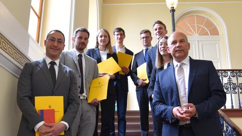 Justizminister Poseck mit sieben Personen, die gelbe Urkunden in den Händen halten, auf einer Treppe. Alle schauen in die Kamera