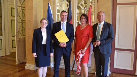 Justizminister Poseck mit Dr. Alexander Seitz, Justizstaatssekretärin Eichner und einer weiteren Frau in einem Saal vor Flaggen. Dr. Seitz hält die Ernennungsurkunde in Händen, alle lächeln in die Kamera.