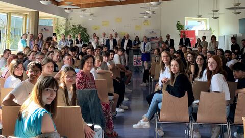 Schülerinnen und Schüler in einem Saal sitzen auf Stühlen verteilt im Raum und drehen sich zur Kamera