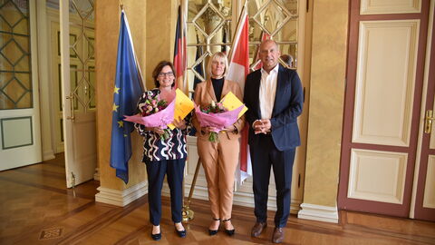 Justizminister Poseck mit der neuen Leiterin der JVA Limburg Carmen Röhrig und der bisherigen Leiterin Anja Müller. Sie stehen gemeinsam mit den Urkunden in Händen vor einigen Flaggen (Europa, Deutschland und Hessen).
