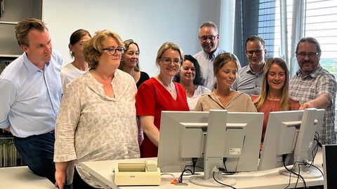 Justizstaatssekretärin Eichner steht mit mehreren Personen an einem Schriebtisch vor zwei Bildschirmen.