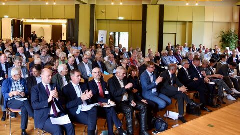 Blick auf einen mit Menschen gefüllten Saal. Die Personen sitzen auf Stühlen einem Podium zugewandt.