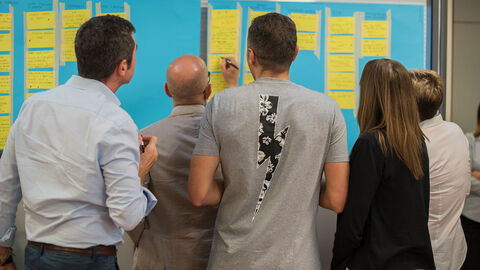 Ideensammlung (Symbolbild) Mehrere Personen stehen vor einem Ständer, an dem gelbe Haftnotizzettel angeklebt sind.