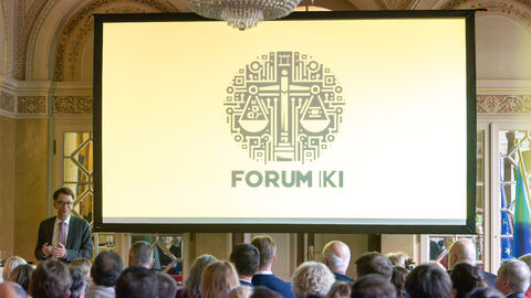 Eine große Leinwand mit einer Grafik und dem Titel "Forum KI" hängt vor einem Publikum, das dieser zugewandt sitzt.