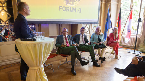 Mehrere Personen sitzen unterhalb einer Leinwand auf der "Forum KI" zu lesen ist und sind einem Publikum zugewandt. Am linken Rand steht eine Person an einem Stehtisch und moderiert.