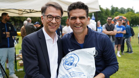 Justizminister Heinz steht neben einer weiteren Person auf einer grünen Wiese. Sie halten einen weißen Beutel zwischen sich. Im Hintergrund sind weitere Personen zu sehen.