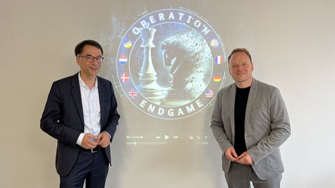 Justizminister Heinz steht vor Wand, auf der mittels eines Beamers das Logo "Operation Endgame" zu sehen isr