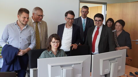 Besuch Digitaler Servicepoint Justizminister Heinz steht mit einer Gruppe weiterer Personen um einen Schreibtisch, an dem eine Person arbeitet. Sie schauen alle auf die Bildschirme.