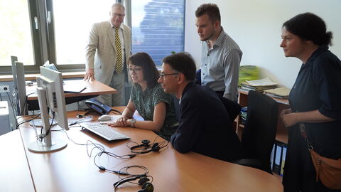 Digitaler Servicepoint Justizminister Heinz sitzt neben einer Dame an einem Schreibtisch. Sie schauen gemeinsam auf die Monitore vor ihnen. Weitere Personen stehen hinter Ihnen und sehen ebenfalls zu.
