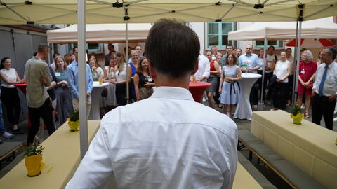 Justizminister Heinz ist von hinten zu sehen. Es steht in einem Hof wo mehrere Pavillons stehen und Menschen um ihn stehen.