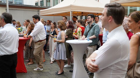 Menschen stehen versammelt in einem Innenhof und schauen nach links, wo sie einer Rede zuhören.