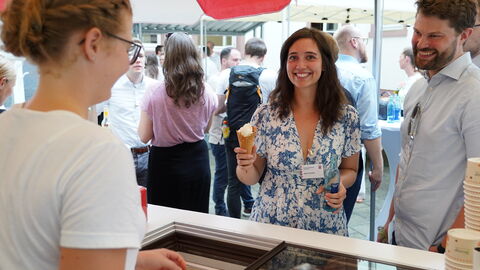 Eine Frau und ein Mann stehen an einem Stand an, wo sie sich ein Eis kaufen. Die Frau hält eins bereits in der Hand und lacht.