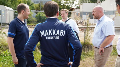 Justizminister Heinz besucht TuS Makkabi Frankfurt e. V. Mehrere Personen im Gespräch mit Justizminister Heinz. Ihn sieht man von hinten mit einer Sportjacke, auf der "Makkabi Frankfurt" steht.