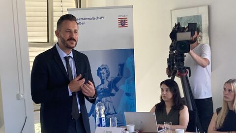 Staatsanwaltschaft Gießen Ein Mann in Anzug steht im Raum vor Personen, die ihm zuschauen und erzählt etwas. Im Hintergrund ein Kameramann und ein Aufsteller.