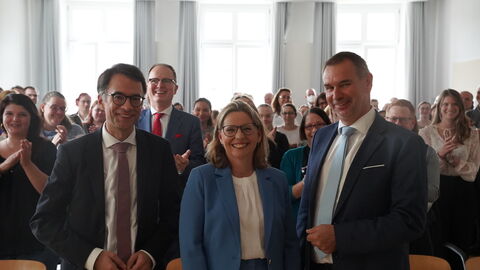 3 Personen stehen nebeneinander und lächeln in die Kamera. Hinter ihnen stehen weitere Menschen.