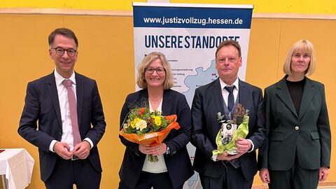 4 Personen stehen nebeneinander in einer Turnhalle. Die Personen in der Mitte halten Blumen und ein Präsent in der Hand.