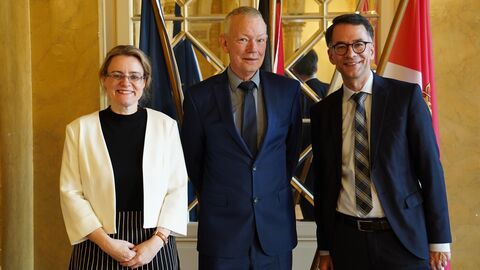 Ruhestand Thomas Puffert  Zwei Männer und eine Frau lachen in Kamera. Im Hintergrund sieht man drei Flaggen.