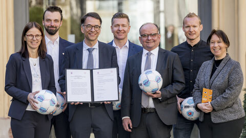 Justizminister Heinz steht mit weiteren Personen zusammen. Er hält ein Dokument in Händen, die anderen Personen halten Fußbälle.