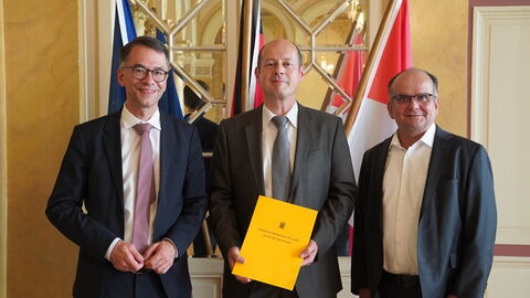 Ulf Frenkler Drei Männer stehen nebeneinander. Der Mann in der Mitte hält ein Dokument in der Hand.