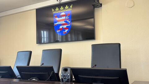 Drei Stühle hinter einem Tisch. Vor jedem Stuhl ist ein Bildschirm. Hinter den Sitzen hängt ein Bildschirm mit dem Hessischen Wappen.