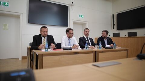 Vier Personen sitzen an einem Tisch in einem Saal.