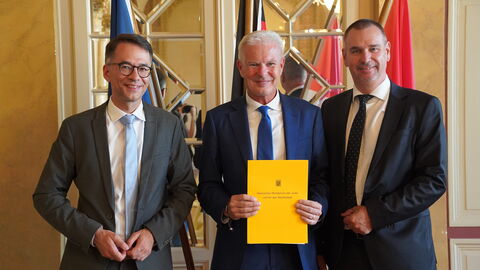 Drei Männer im Anzug lachen in Kamera. Hinter ihnen befinden sich Flaggen.