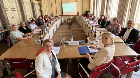 Mehr als 20 Personen sitzen an Tischen, die in einem U aufgestellt worden. Sie sitzen in einem großen Saal.