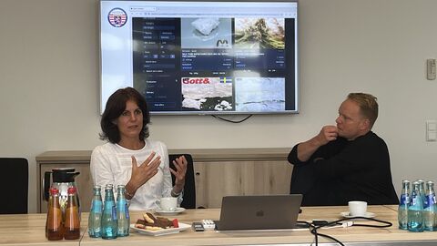 Zwei Personen sitzen an einem Tisch. Im Hintergrund ist ein Bildschirm mit einer Präsentation.