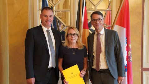 Drei Personen stehen vor einer Glastür nebeneinander. Die Frau in der Mitte hält ein Dokument in der Hand.