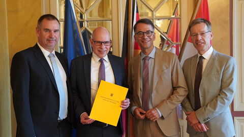 Vier Männer stehen im Anzug vor 3 Flaggen. 