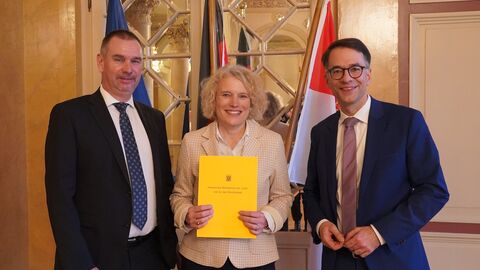 Zwei Männer und eine Frau stehen vor Flaggen von Europa, Deutschland und Hessen. Die Dame hält eine Urkunde in den Händen.