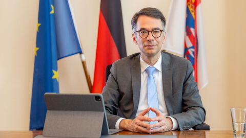 Justizminister Hein sitzt an einem Tisch. Neben ihm steht ein Tablet und im Hintergrund sind die Flaggen von Europa, Deutschland und Hessen aufgestellt.