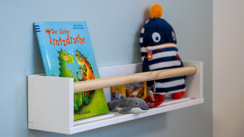 Regal, das an der Wand hängt. Darauf ist ein Buch und Kuscheltiere platziert.