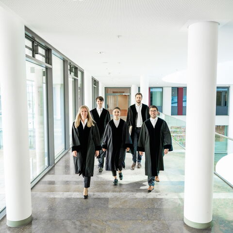 Zwei Frauen und ein Mann in Richterroben laufen in einem Gang entlang auf die Kamera zu, links und rechts des Ganges stehen zwei große weiße Säulen.